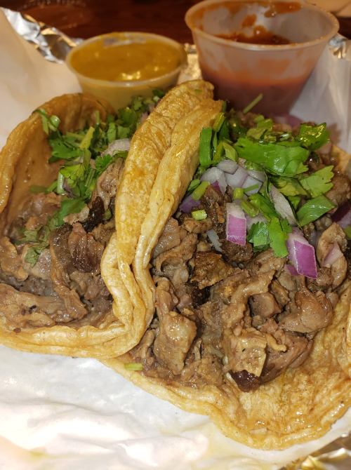 Two tacos filled with seasoned meat, topped with chopped onions and cilantro, served with salsa and guacamole on the side.
