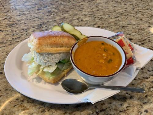 A plate with a sandwich, a bowl of soup, pickles, and a packet of crackers on a napkin.