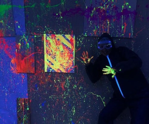 A person in a black outfit and goggles poses against a brightly splattered wall in neon colors.