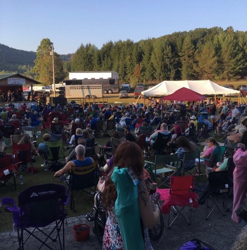 A large crowd sits in folding chairs at an outdoor event, with tents and a stage in the background, surrounded by trees.