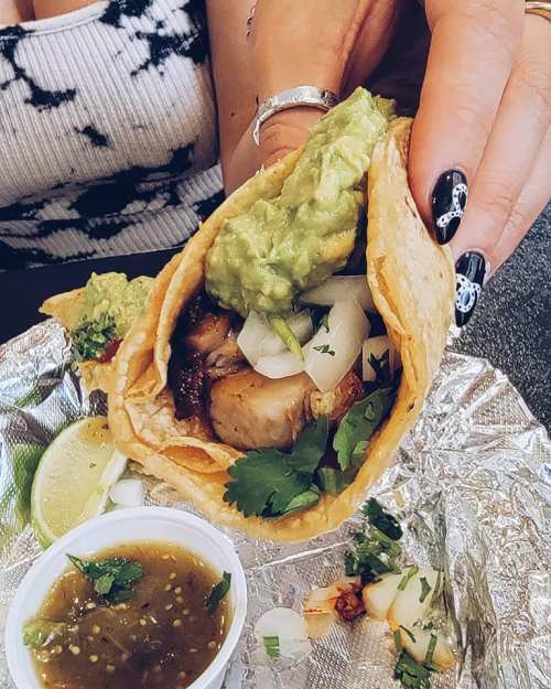 A hand holding a taco filled with meat, guacamole, onions, and cilantro, alongside a small bowl of green salsa and lime.