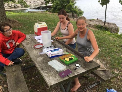 Three people sit at a picnic table by a lake, working with scientific equipment and notebooks.