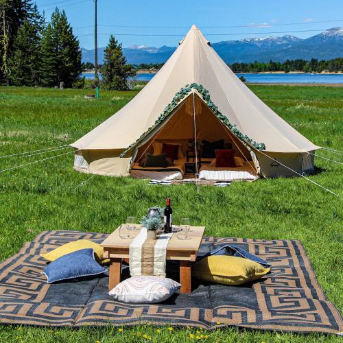 A spacious tent set up in a grassy area, with a picnic table and cushions nearby, surrounded by mountains and trees.