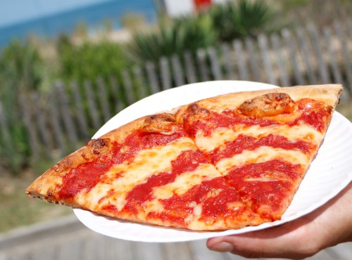 A hand holding two slices of pizza with cheese and tomato sauce, set against a beach background.