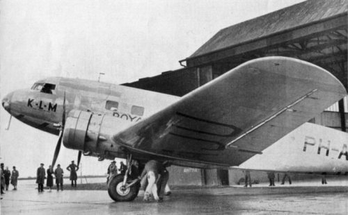 A vintage airplane with KLM markings parked in front of a hangar, surrounded by people on a rainy day.