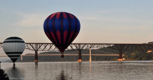 Two hot air balloons float over a river, with a bridge in the background and a serene sunset sky.