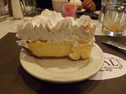 A slice of lemon pie topped with whipped cream on a white plate, set on a dark tablecloth.