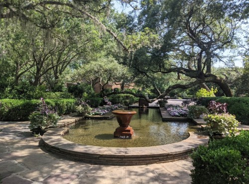 A serene garden with a central fountain, surrounded by lush greenery and flowering plants under a clear blue sky.