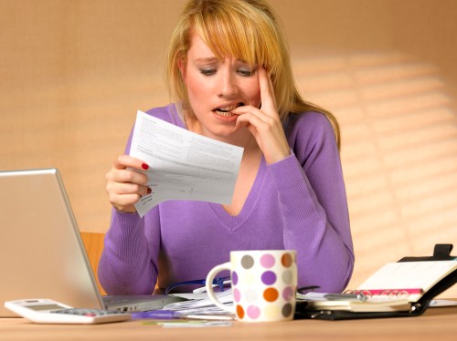 A worried woman in a purple sweater looks at a document while sitting at a desk with a laptop and a coffee mug.
