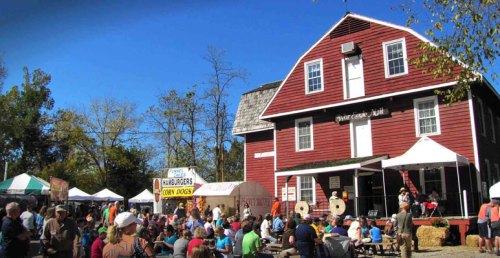 A lively outdoor festival scene with a red barn, crowds of people, food stalls, and a clear blue sky.