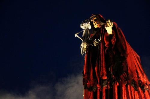 A tall figure in a red cloak and skull mask stands against a dark sky, holding a skeletal puppet.