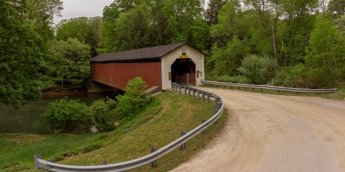 A red covered bridge spans a calm river, surrounded by lush green trees and a winding dirt road.