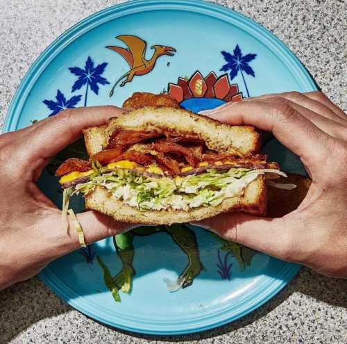 A person holds a sandwich filled with bacon, lettuce, and egg, on a colorful plate with a tropical design.