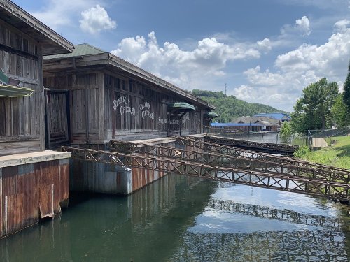 Abandoned wooden buildings by a calm waterway, with a bridge connecting to the shore under a blue sky.