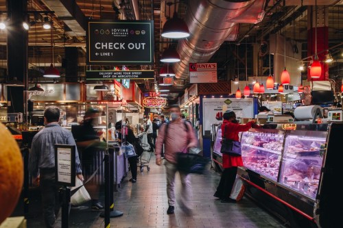 A bustling market scene with shoppers, food stalls, and a "CHECK OUT" sign overhead. Bright lights and vibrant activity.