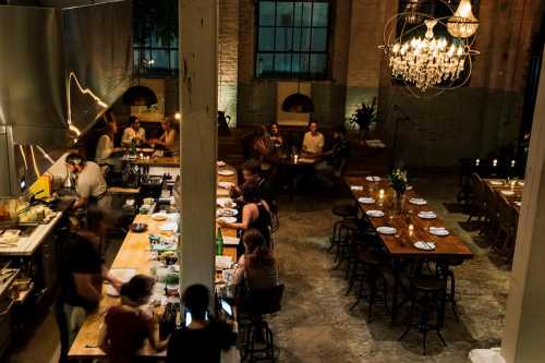 A cozy restaurant interior with diners at tables, a bustling kitchen, and a chandelier overhead. Warm, inviting atmosphere.