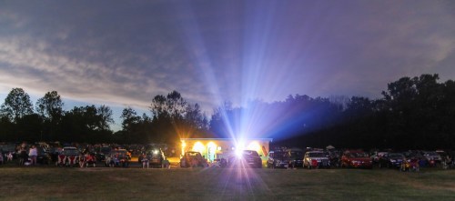 A drive-in movie scene at dusk, with cars parked in a field and a bright screen illuminating the night sky.