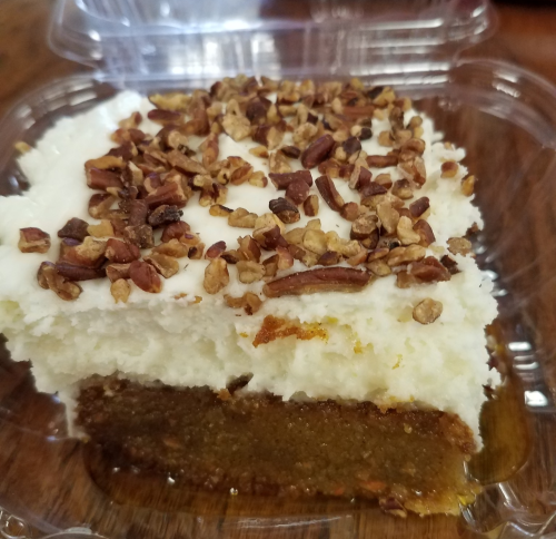 A close-up of a dessert slice with cream frosting and chopped nuts on top, in a clear plastic container.
