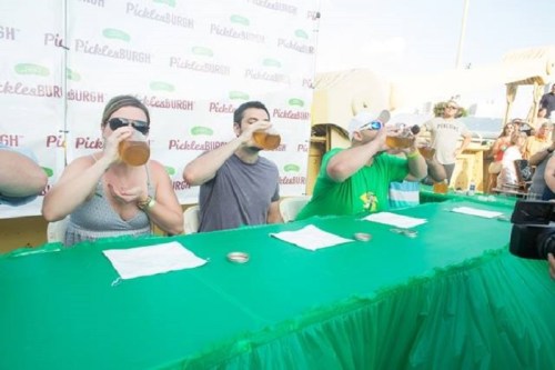 Three people drinking from large glasses at a festive outdoor event with a green table and a crowd in the background.