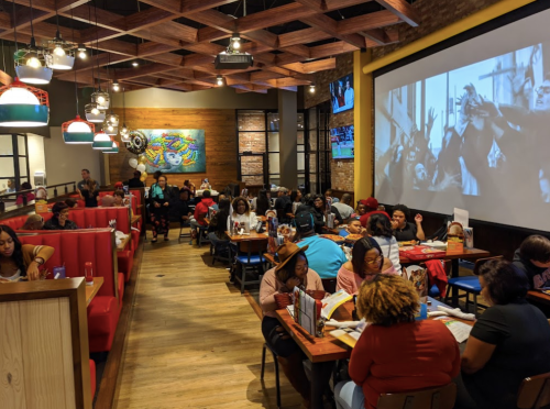 A busy restaurant interior with diners at tables, colorful decor, and a large screen showing a film in the background.