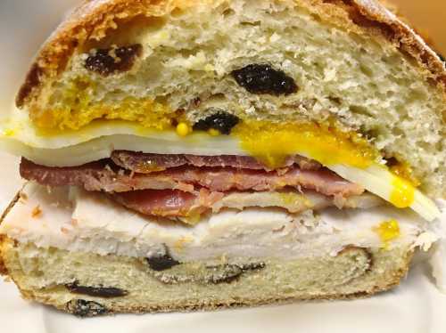 Close-up of a sandwich with layers of turkey, ham, cheese, and egg on a sliced, raisin-studded bread.