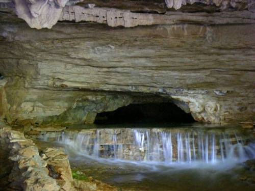 A serene underground cave with a small waterfall flowing into a dark pool, surrounded by rocky formations.