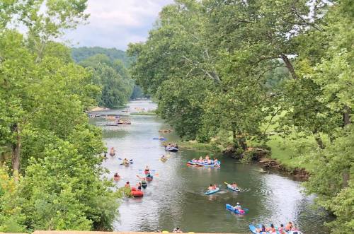 A scenic river with people kayaking and canoeing, surrounded by lush green trees and a cloudy sky.