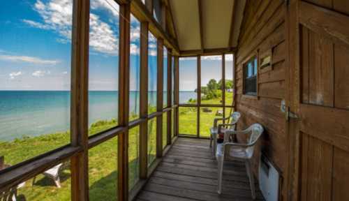 A screened porch with two chairs overlooking a serene lake and green landscape under a blue sky.