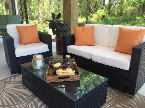 Cozy outdoor seating area with orange cushions, a glass coffee table, and a tray of snacks surrounded by greenery.