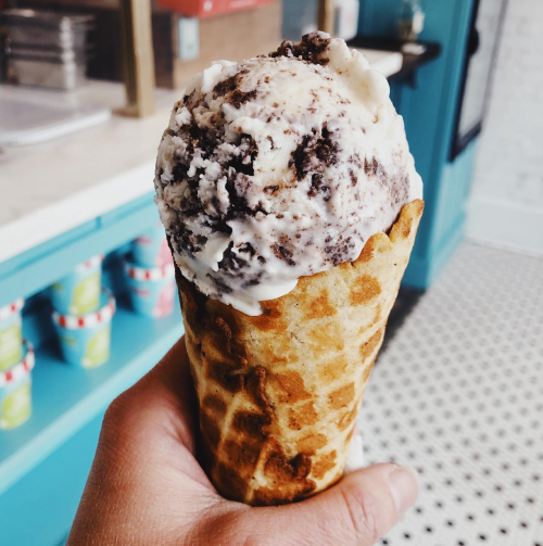 A hand holding a waffle cone filled with cookies and cream ice cream against a colorful background.