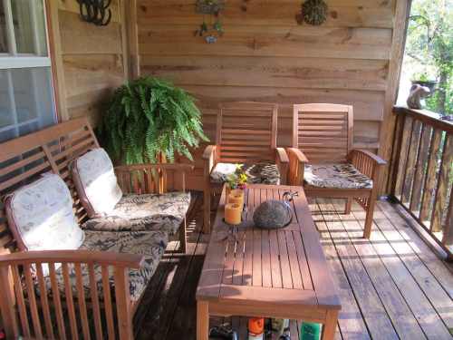 A cozy wooden porch with two chairs and a sofa, adorned with cushions, and a table with a potted plant and a rock.