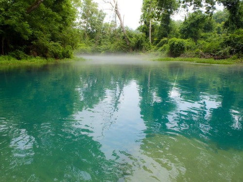 A serene, misty lake surrounded by lush greenery, reflecting the vibrant blue sky and trees.
