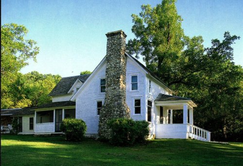 A charming white house with a stone chimney, surrounded by lush greenery and trees on a sunny day.