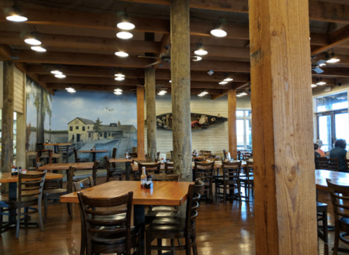 Interior of a rustic restaurant with wooden beams, tables, and a mural of a waterfront scene on the wall.