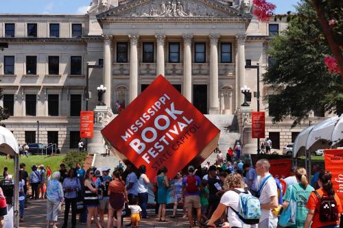 Crowd gathered at the Mississippi Book Festival outside the State Capitol, featuring a large orange cube sign.