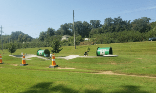 A grassy park with green tunnels and orange traffic cones, surrounded by trees and a clear blue sky.