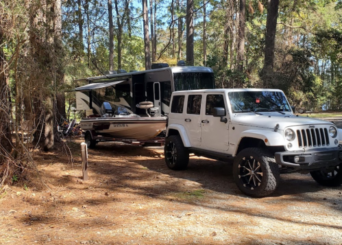 A white Jeep parked next to a camper trailer and a boat in a wooded area. Pine trees surround the scene.