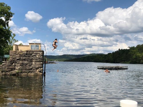 A person jumps off a stone platform into a lake, with clouds and trees in the background.