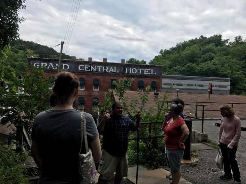 A group of people stands near the Grand Central Hotel, surrounded by greenery and a cloudy sky.