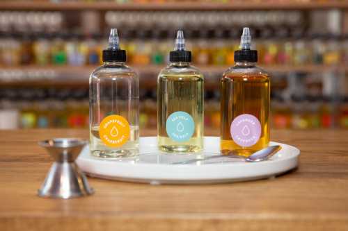 Three bottles of flavored oils on a white tray, with a jigger and a spoon in the foreground, set against a colorful background.