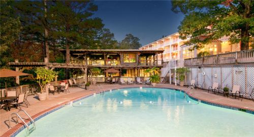 A serene pool area with a wooden deck, surrounded by trees and a hotel in the background, illuminated at dusk.