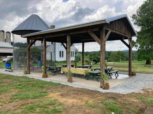 A wooden pavilion with picnic tables, surrounded by grass, near a farm building and silos under a cloudy sky.