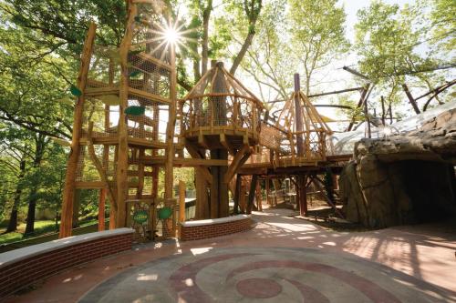 A wooden playground structure with towers and slides, surrounded by trees and sunlight filtering through the leaves.