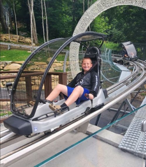 A child smiles while riding a mountain coaster on a track surrounded by trees.