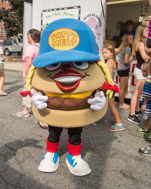 A cartoonish burger mascot named Patty Melt, wearing a blue cap and red shoes, stands in a crowd at a fair.