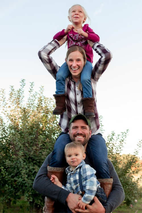A happy family poses outdoors, with two children on top of a smiling parent, surrounded by trees.