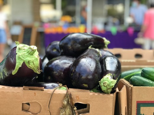 Fresh eggplants stacked in cardboard boxes at a market, with blurred shoppers and colorful decorations in the background.