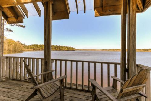 View from a wooden porch overlooking a calm lake surrounded by trees under a clear blue sky.