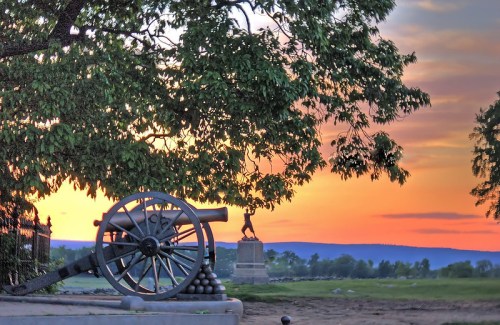 A historic cannon beside a tree, with a sunset backdrop and a statue in the distance.
