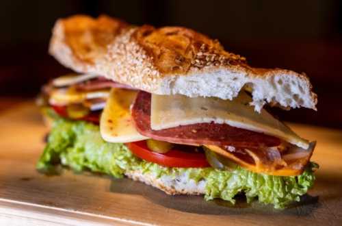 A close-up of a sandwich with layers of lettuce, tomato, cheese, salami, and pickles on a sesame seed bun.